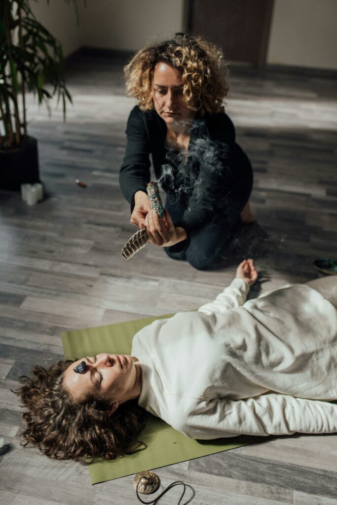 A woman performs a healing ritual with smudging and crystals on another woman indoors.