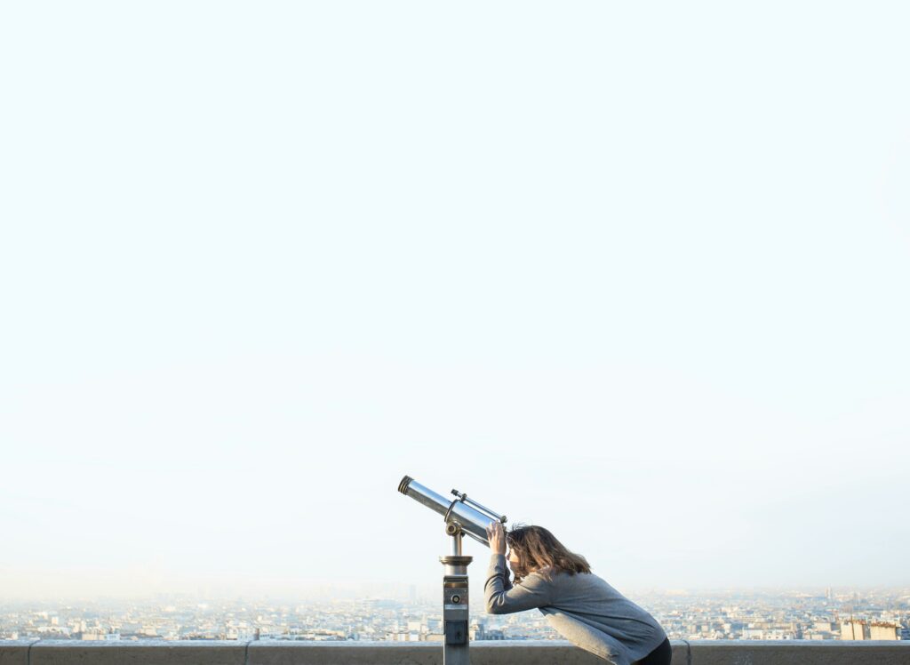 A woman peers through a coin-operated telescope overlooking a city skyline.